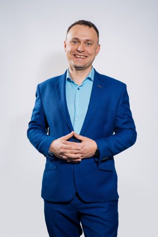 Mężczyzna ubrany w niebieski garnitur i jasnoniebieską koszulę stoi uśmiechnięty na prostym, jasnym tle, z rękami luźno splecionymi przed sobą - promieniując pewnością siebie często spotykaną u profesjonalistów UX Design.