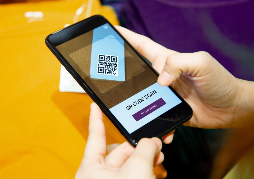 Osoba trzyma smartfon, skanując kod QR na etykiecie produktu. Ekran telefonu wyświetla interfejs skanowania kodu QR z przyciskiem oznaczonym SZCZEGÓŁY PRODUKTU. Scena sugeruje mobilne zakupy lub dostęp do informacji o produkcie.