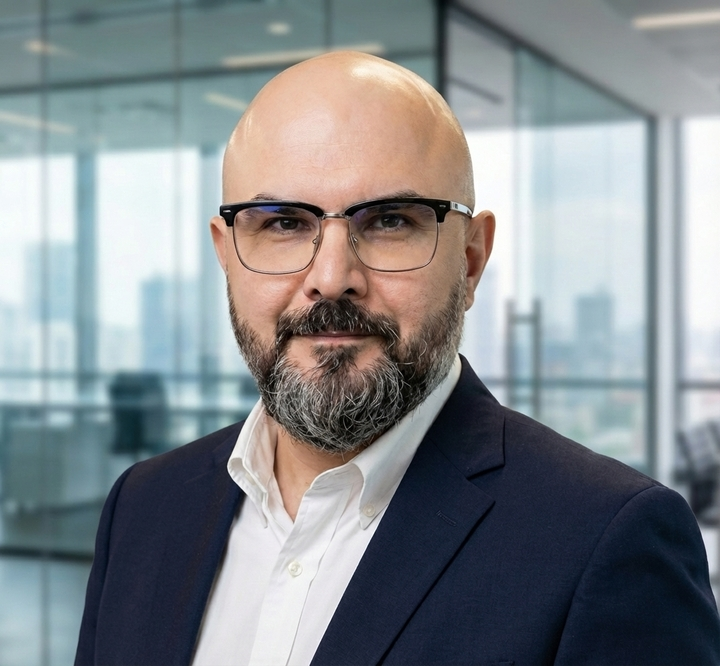 A bald man with glasses and a beard, wearing a navy suit and white shirt, stands in a modern office with large glass windows and a city view in the background.