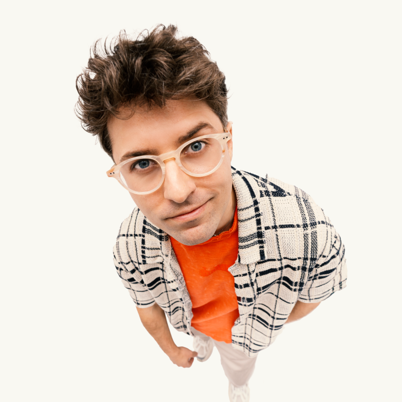 Młody mężczyzna z falującymi brązowymi włosami, w przezroczystych okularach, pomarańczowej koszuli i czarno-białej kraciastej kurtce patrzy w kamerę, stojąc na białym tle.