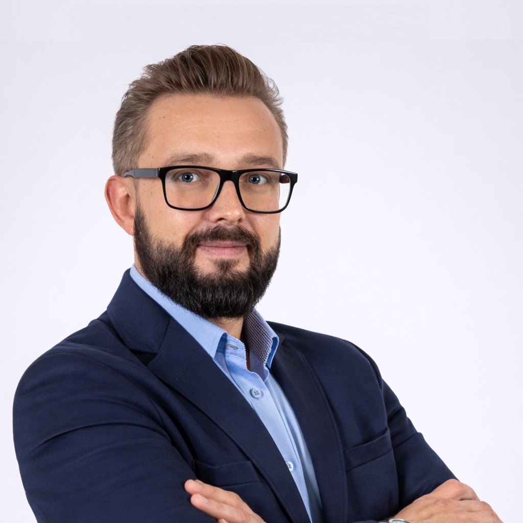 Mężczyzna w okularach, z starannie przystrzyżoną brodą i krótkimi włosami ma na sobie granatową marynarkę i jasnoniebieską koszulę. Stoi na prostym białym tle z założonymi rękami, patrząc pewnie w obiektyw — gotowy, by podzielić się swoimi spostrzeżeniami na temat sztucznej inteligencji w handlu detalicznym podczas zbliżających się spotkań Community Talks.