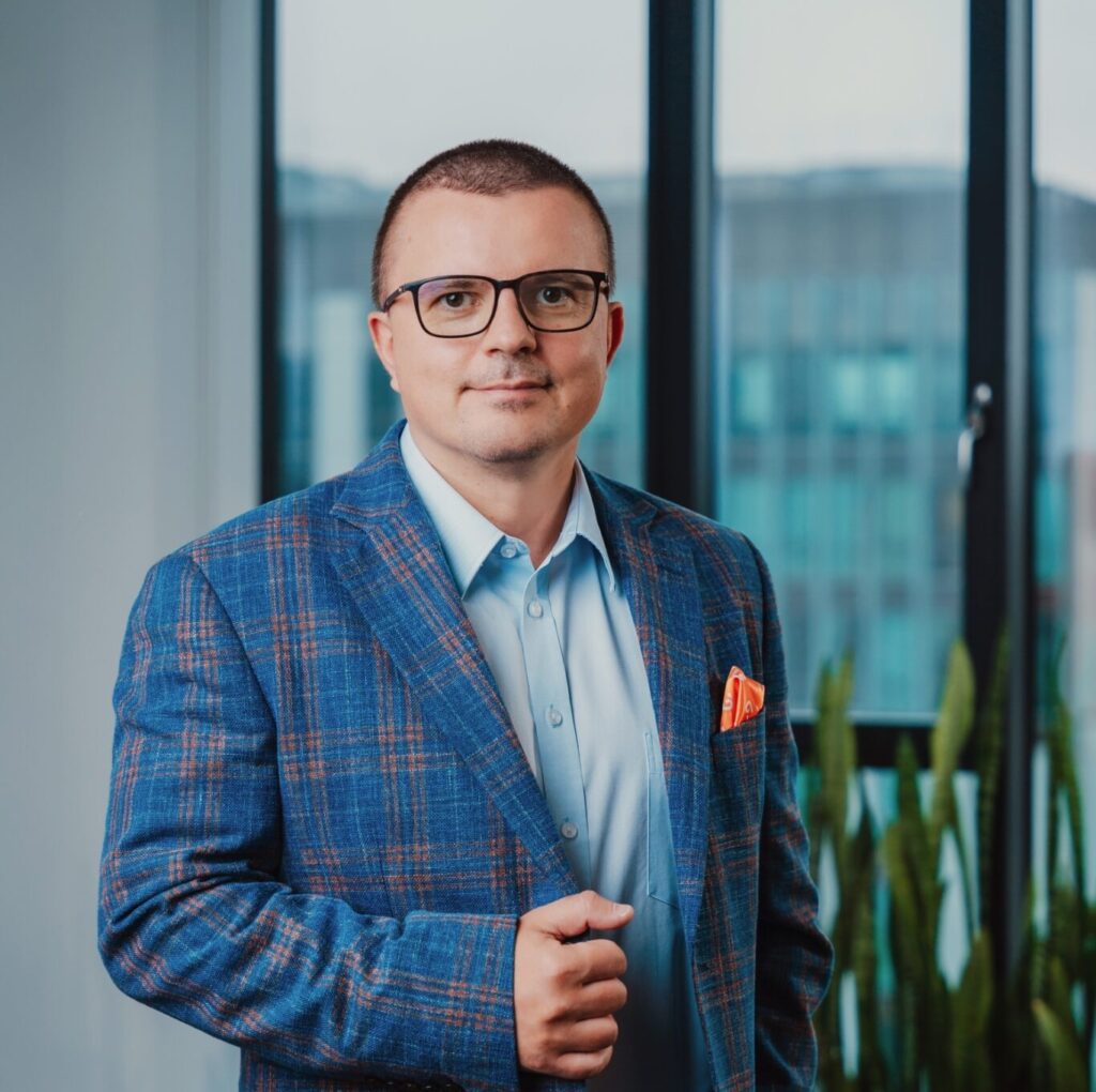 Mężczyzna w okularach i niebieskiej marynarce w kratę z pomarańczową poszetką stoi w pomieszczeniu przed dużymi oknami, a obok niego widać zielone rośliny — gotowy, by podzielić się swoją wiedzą na temat sztucznej inteligencji i spotkań społecznościowych w ramach środowiska detalicznego „Ścieżka klienta”.