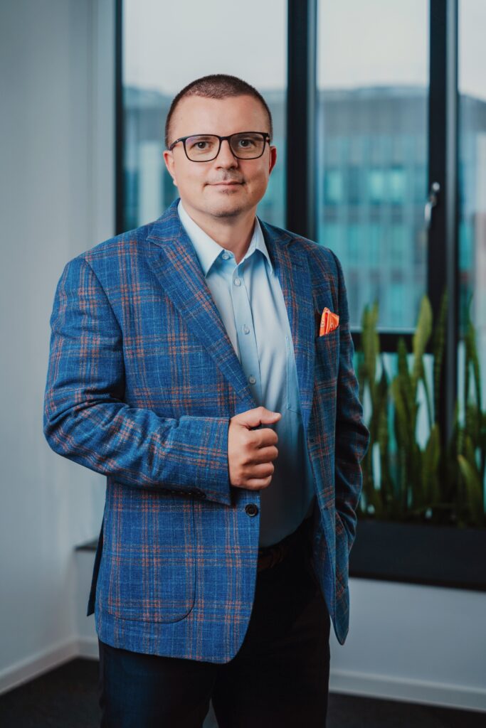 A man wearing glasses, a blue plaid blazer, and a light blue shirt stands indoors by a window with city buildings outside and tall green plants nearby.