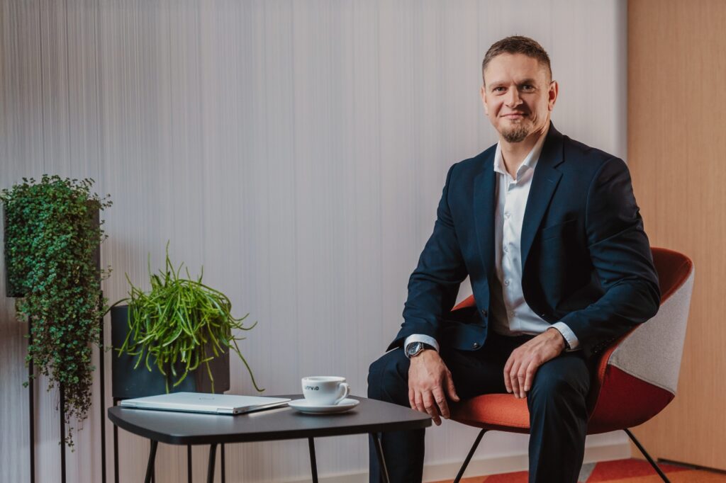 Mężczyzna w ciemnym garniturze siedzi na nowoczesnym krześle z uśmiechem na twarzy; na małym stoliku stoi laptop i filiżanka kawy – być może analizuje strategie mające na celu zwiększenie przychodów. Tło zdobią dwie zielone rośliny.