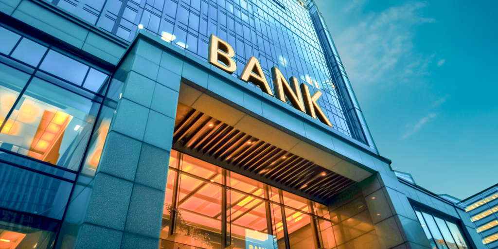 Nowoczesny budynek banku z dużymi szklanymi oknami, złotym napisem „BANK” nad wejściem i ciepłym oświetleniem wewnątrz, zaprojektowanym z myślą o optymalizacji konwersji, na tle błękitnego nieba.