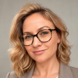 Kobieta o falujących blond włosach, w okularach w oprawkach z szylkretu i jasnej marynarce, uśmiecha się delikatnie do aparatu na neutralnym tle. Ten portret oddaje przyjazną i profesjonalną postawę Katarzyny Zajchowskiej.