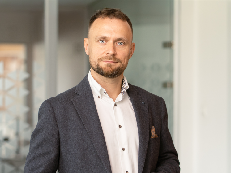 Mężczyzna o krótkich brązowych włosach i brodzie, ubrany w ciemną marynarkę na białą koszulę, stoi w nowoczesnym biurze ze szklanymi ścianami — w otoczeniu idealnym do omawiania strategii takich jak optymalizacja konwersji.