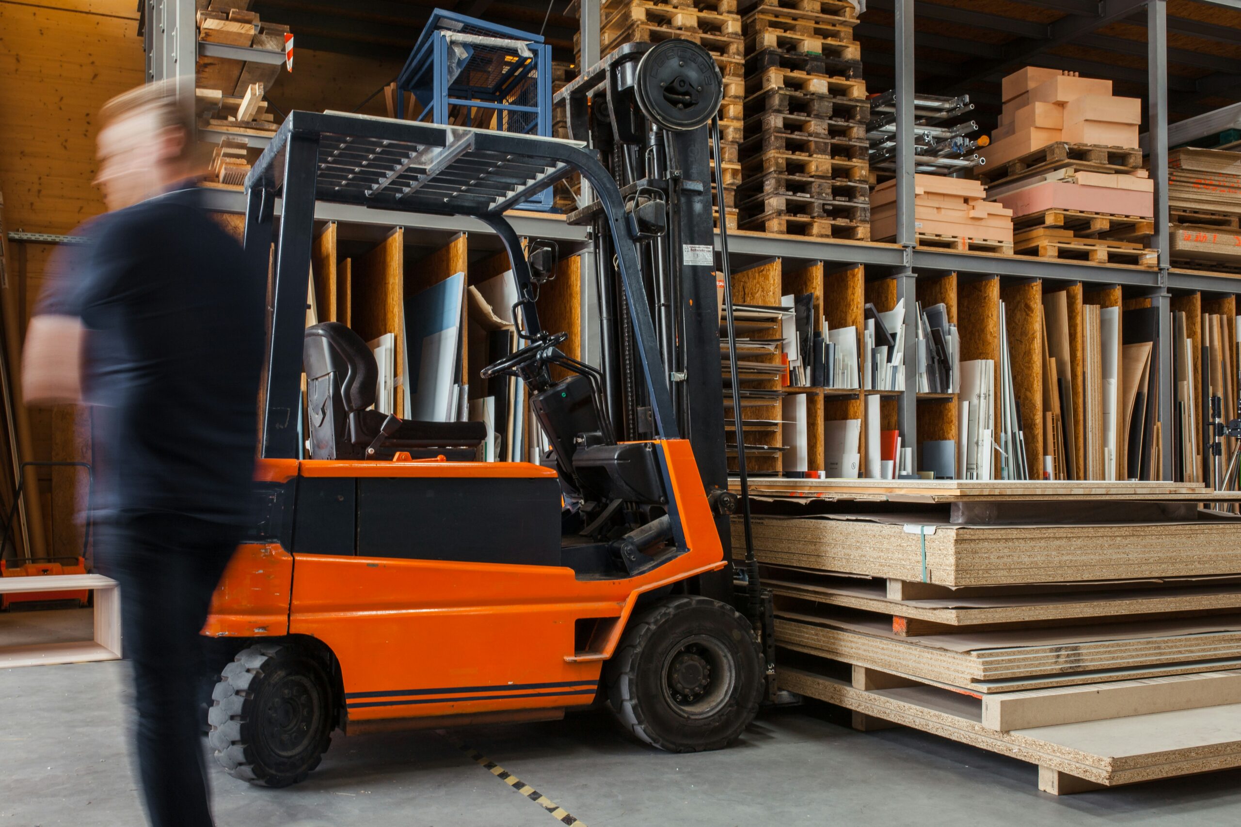 In einem Lager der Wego Systembaustoffe GmbH steht ein orangefarbener Gabelstapler neben Holzstapeln. Eine Person in dunkler Kleidung geht vorbei und erscheint aufgrund der Bewegung verschwommen – eine Szene, die das rasante Tempo des B2B-Vertriebs widerspiegelt. In den Regalen im Hintergrund liegen verschiedene Holzbretter und Materialien, was eine Umgebung veranschaulicht, in der die digitale Transformation die Logistik und den Vertrieb weiterhin prägt.