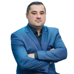 Jerzy Bursztyn, mężczyzna o krótkich ciemnych włosach, ubrany w niebieską marynarkę i wzorzystą koszulę, stoi ze skrzyżowanymi rękami, patrząc pewnie w obiektyw na tle gładkiej białej ściany.
